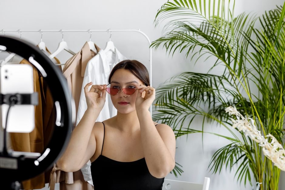Stylish woman wearing sunglasses recording fashion content with a ring light indoors.